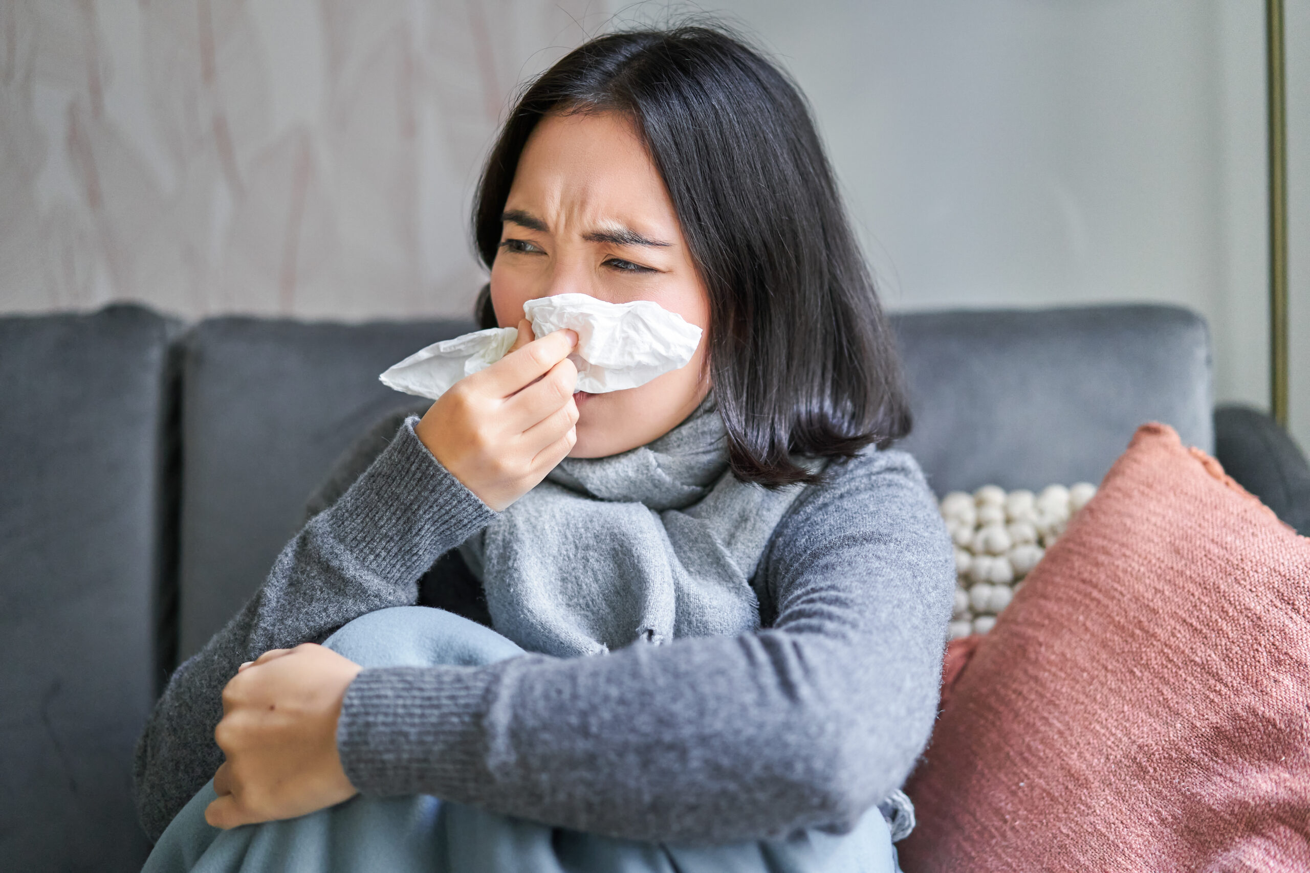 Close up of korean girl sneezing in napking, sitting at home in scarf, feeling unwell, concept of winter, heating cost and people